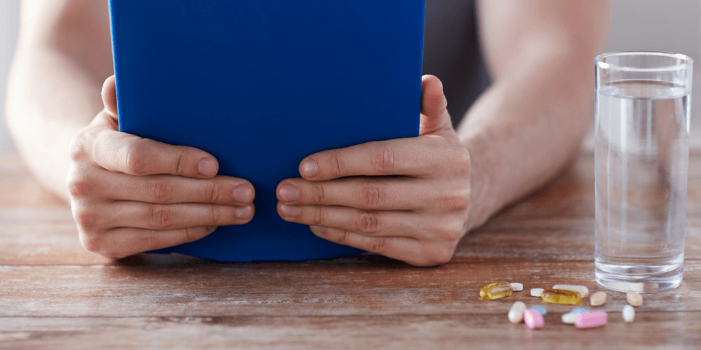 Person holding a blue tablet or book at a wooden table, with a glass of water and a variety of pills and supplements, including capsules and tablets, placed nearby.