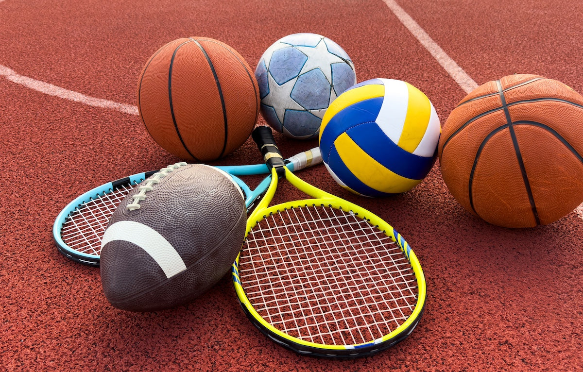 A variety of sports equipment including basketballs, a volleyball, a soccer ball, a football, and two tennis rackets arranged on a red athletic track.