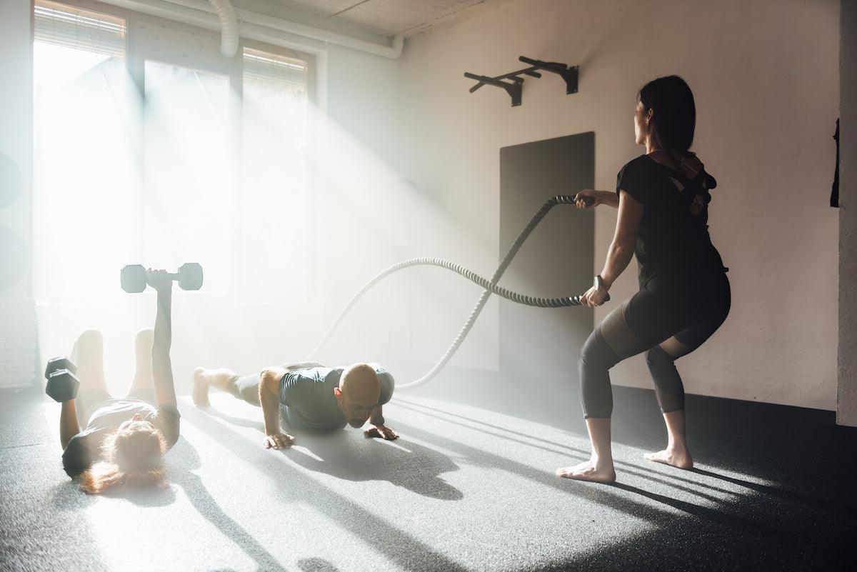 Three people working out in a gym with morning sunlight streaming in—one person using battle ropes, another doing push-ups, and the third lifting dumbbells while lying on their back.