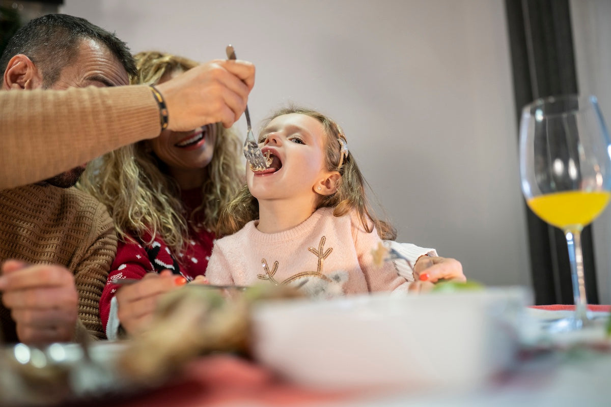 Smiling father feeding his young daughter at the dinner table while the mother watches joyfully in the background, creating a warm and cheerful family mealtime moment.