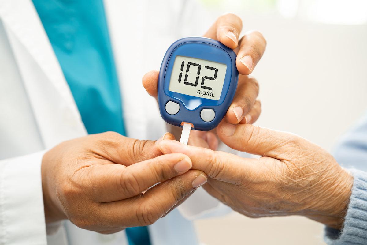 Close-up of a healthcare professional checking a patient's blood sugar level with a glucometer, displaying a reading of 102 mg/dL, highlighting diabetes monitoring and management.