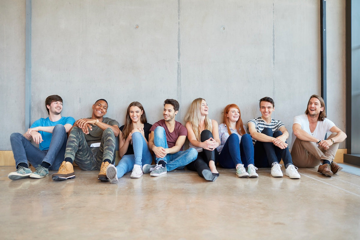 A diverse group of young adults sitting on the floor against a concrete wall, smiling and laughing together in a relaxed, casual setting.
