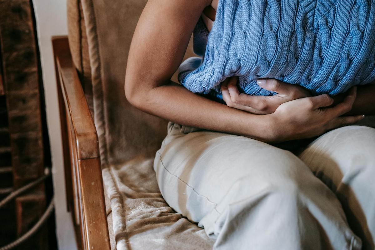 Person sitting on a chair holding their stomach in pain with a blue knitted pillow, suggesting discomfort or gut inflammation.