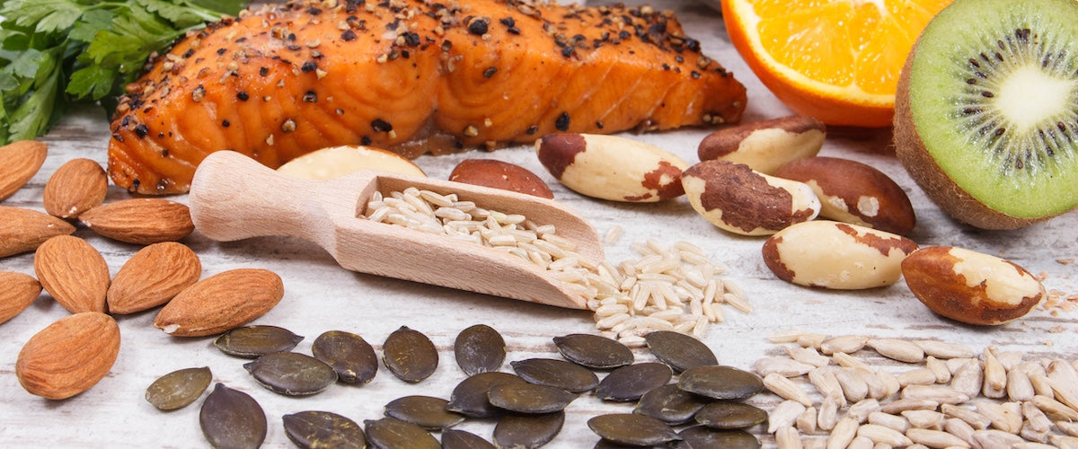 Close-up image of various collagen-boosting foods including grilled salmon, almonds, pumpkin seeds, Brazil nuts, sunflower seeds, brown rice, orange slices, and a halved kiwi, arranged on a rustic wooden surface.
