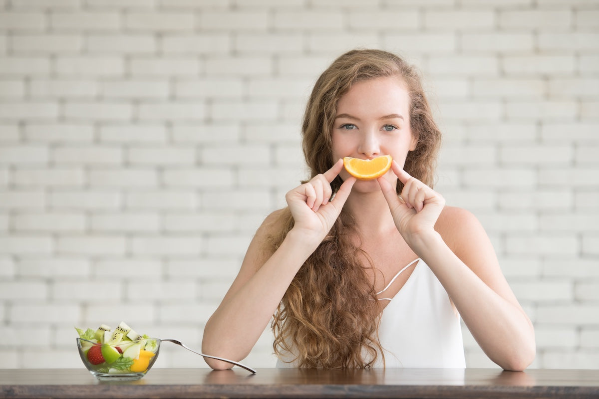 Young woman playfully holding an orange slice like a smile in front of her mouth, sitting at a table with a bowl of fresh fruit, symbolizing the connection between vitamin C and healthy living.