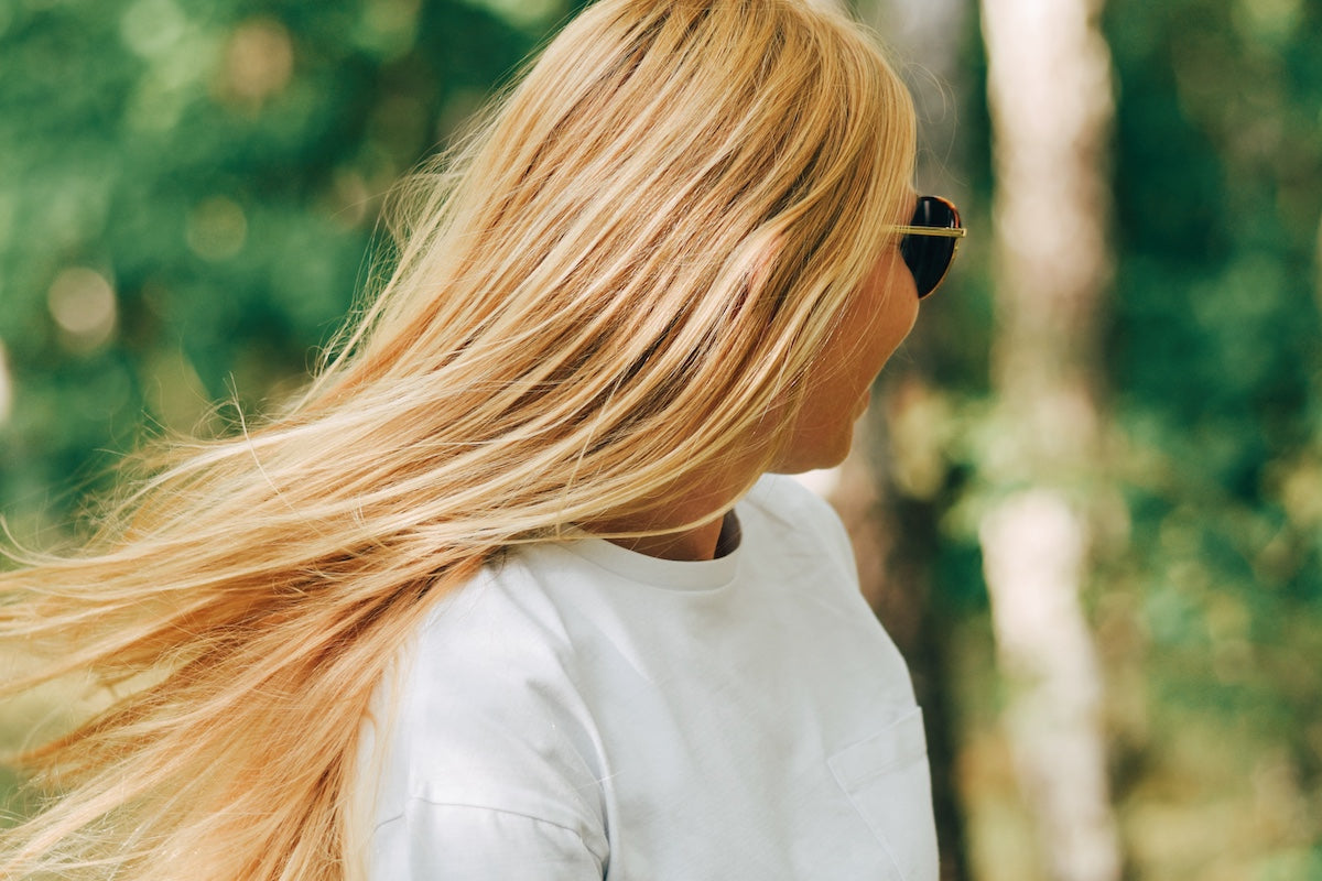 A woman with long, healthy, blonde hair flowing naturally as she turns her head, wearing a white shirt and sunglasses, standing outdoors in a forest with soft, blurred greenery in the background.