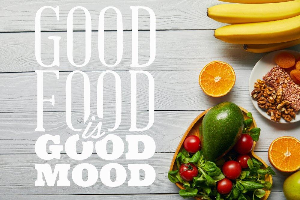 bananas, avocados, oranges, tomatoes, nuts and herbs on white wooden background with "good food is good mood" written on it