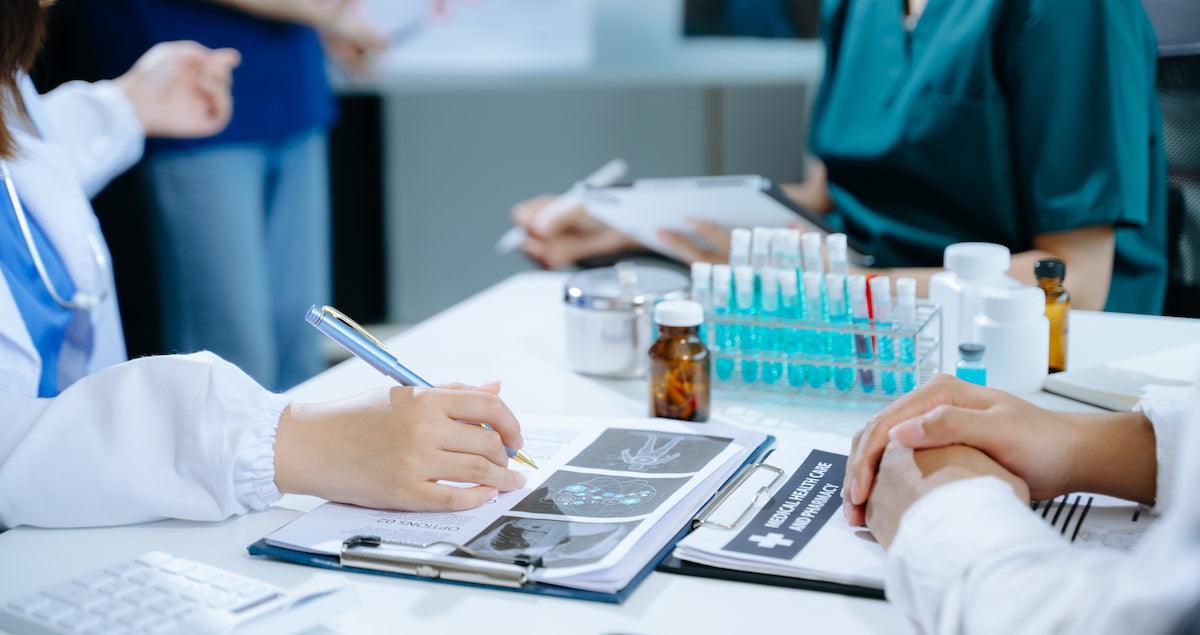 Healthcare professionals discussing medical reports at a desk, with lab samples and medical documents visible. One doctor is writing notes while another reviews data, indicating a collaborative medical analysis setting.
