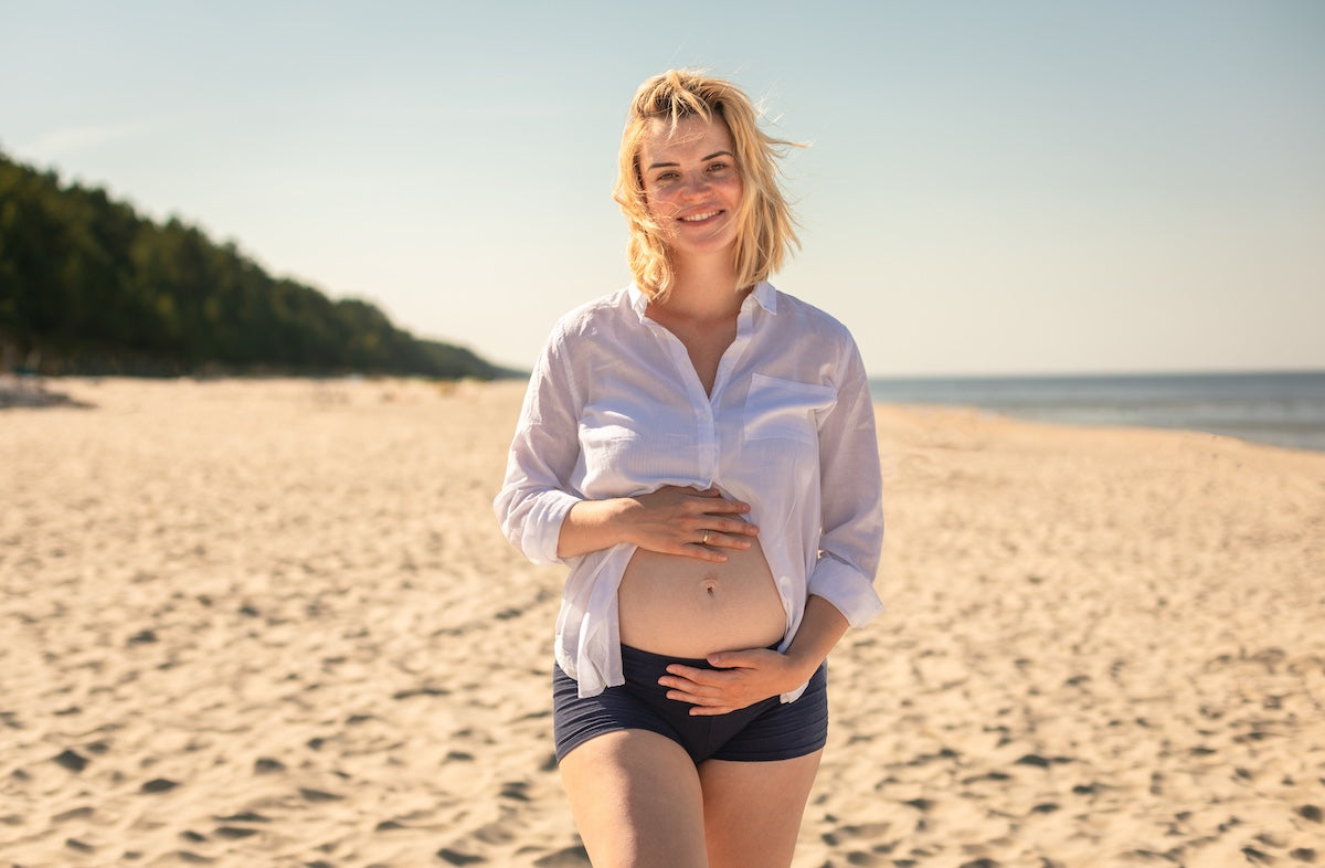 Young pregnant woman in a white shirt and navy shorts smiling gently while standing on a sunny sandy beach, with one hand on her belly and the other below it, surrounded by peaceful seaside scenery.