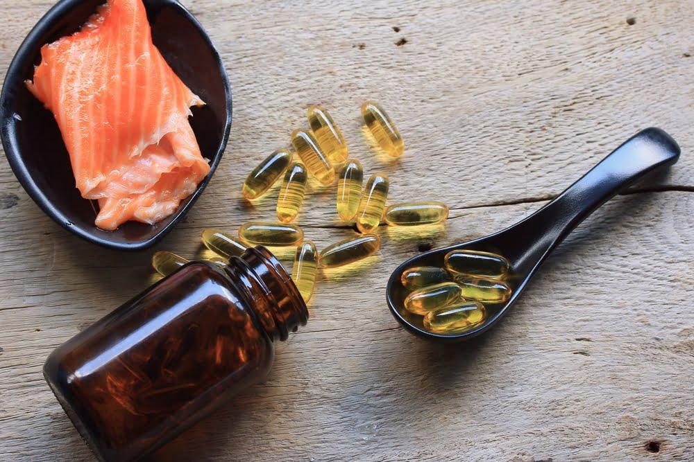A serving of raw salmon, an open brown bottle of omega-3 capsules, and a black spoon filled with capsules arranged on a rustic wooden surface, representing dietary and supplement sources of EPA and DHA.