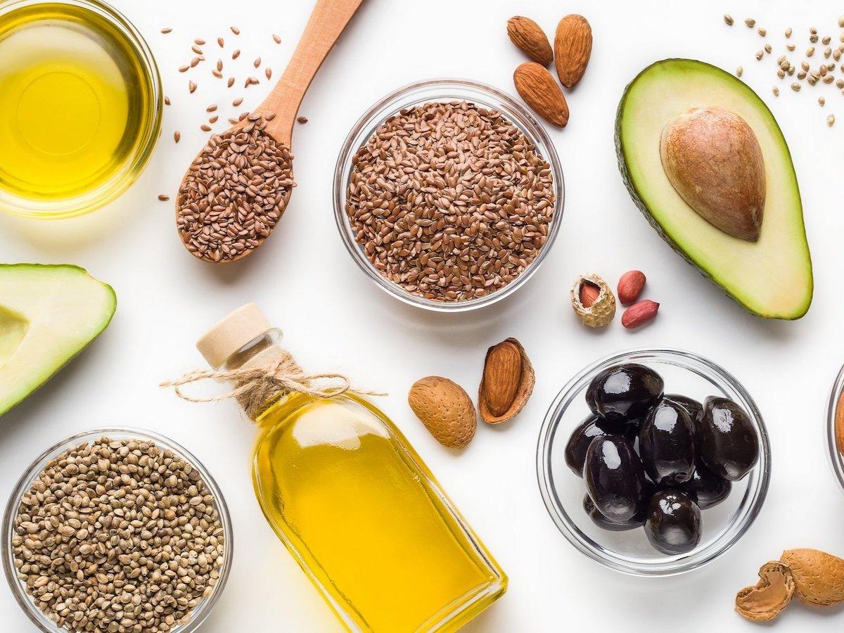 Assorted sources of healthy fats including avocado, flaxseeds, olive oil, hemp seeds, almonds, and black olives, displayed on a white background.