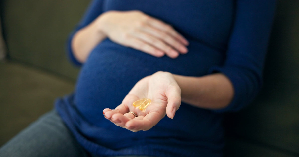Close-up of a pregnant woman sitting on a couch, holding omega-3 or prenatal supplement capsules in one hand while the other hand rests on her belly.