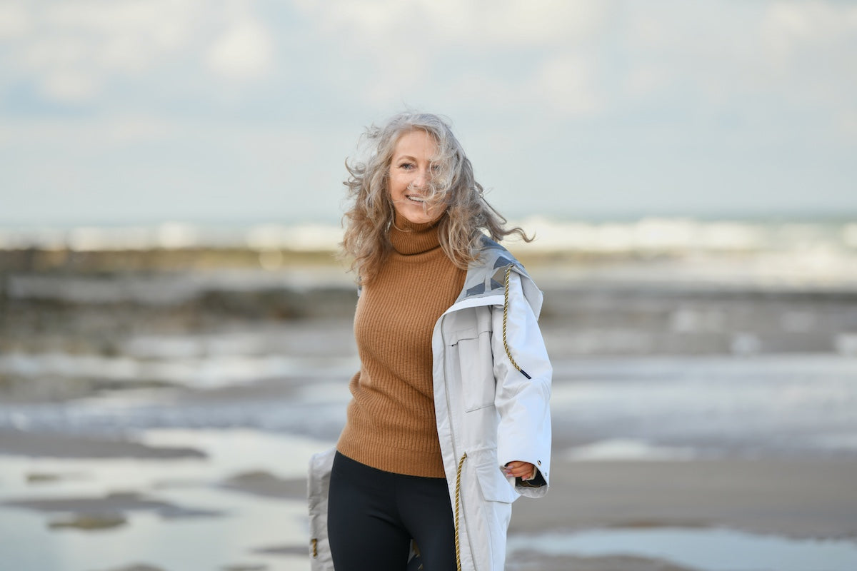 Smiling older woman with curly gray hair wearing a brown sweater and white coat, standing on a beach with the ocean and sky in the background.