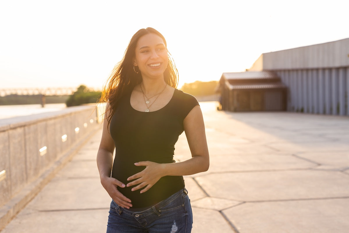 Pregnant woman in casual clothes smiling and touching her belly while walking outdoors at sunset, representing maternal wellness and healthy pregnancy.