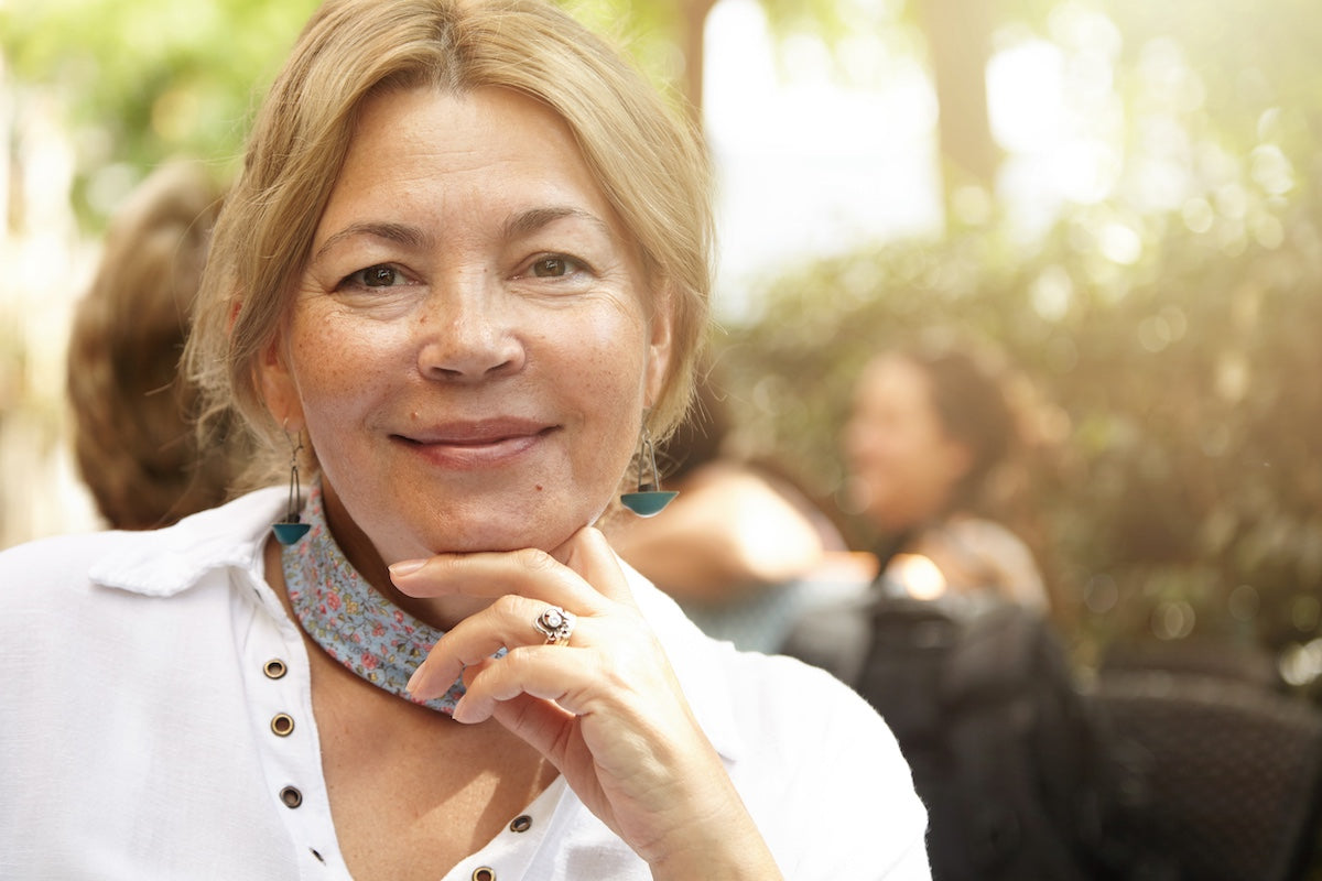 Confident older woman with light brown hair smiling outdoors, wearing a white shirt and floral scarf, with soft natural sunlight in the background.