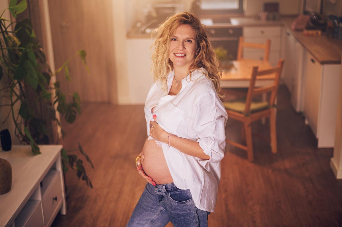 Smiling pregnant woman standing in a cozy home setting, gently holding her baby bump, symbolizing pregnancy and maternal well-being.