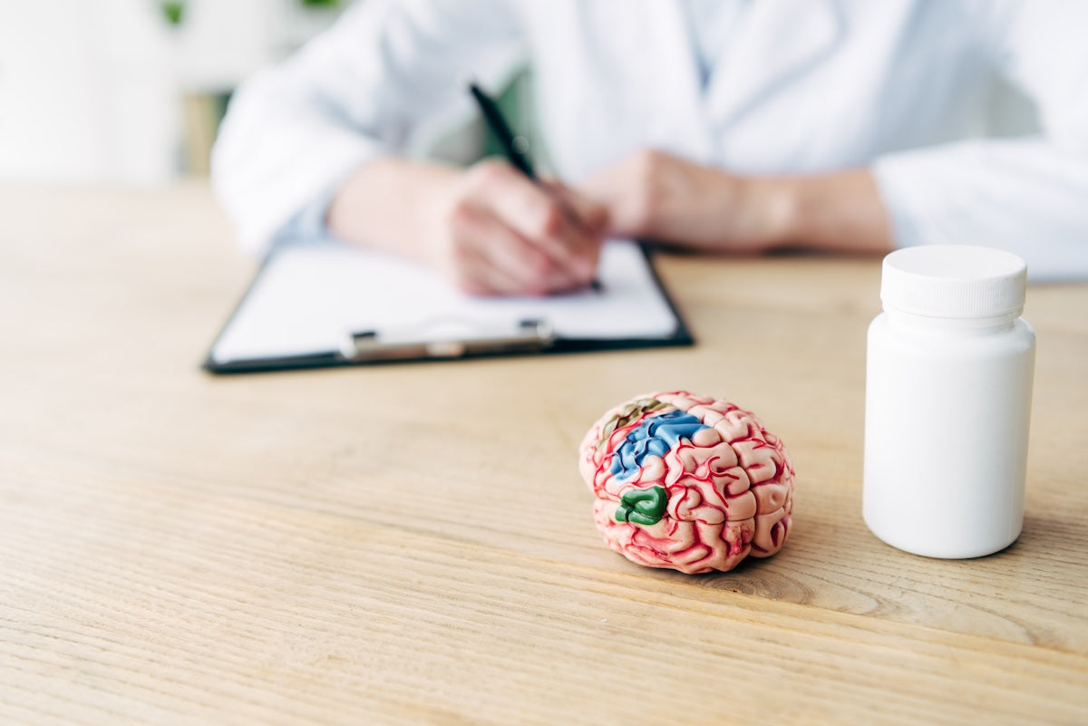 Selective focus of a white pill bottle and a detailed anatomical model of the human brain on a wooden desk, with a doctor writing notes on a clipboard in the blurred background, representing medical consultation or cognitive health treatment.