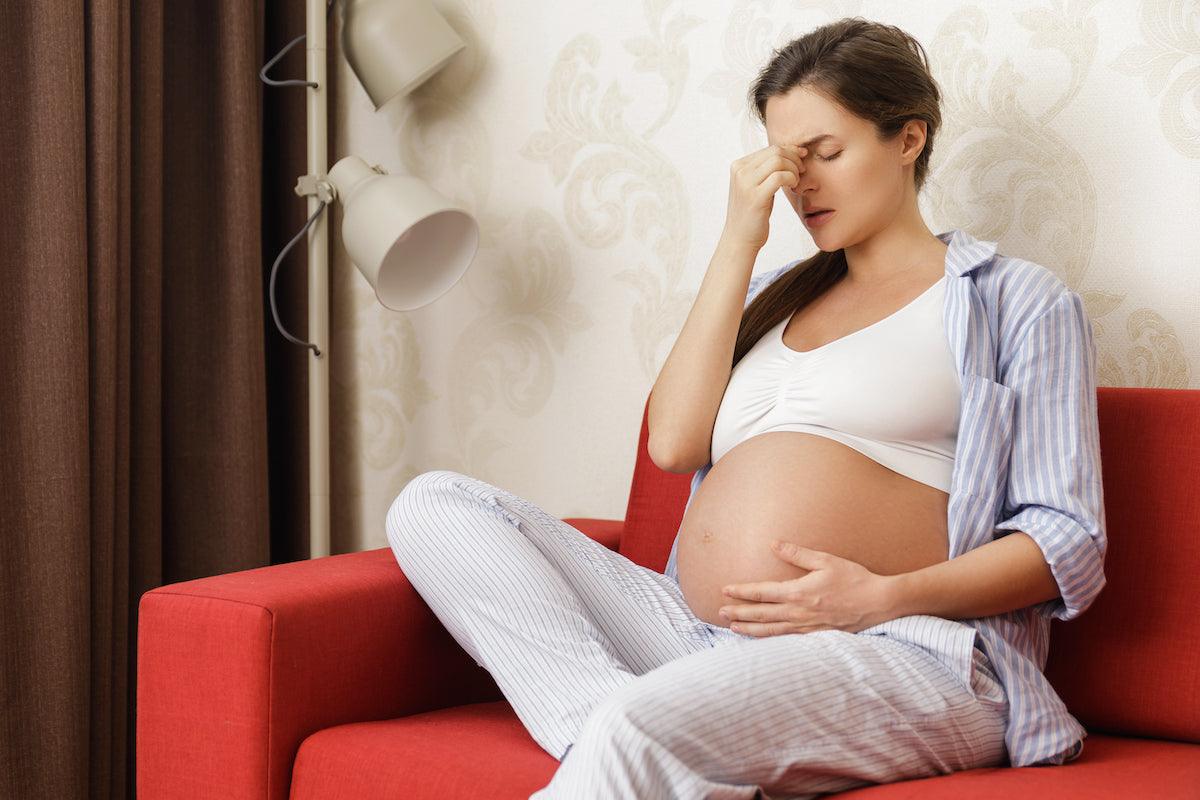 Pregnant woman sitting on a red couch, touching her forehead in discomfort, possibly experiencing stress or fatigue.