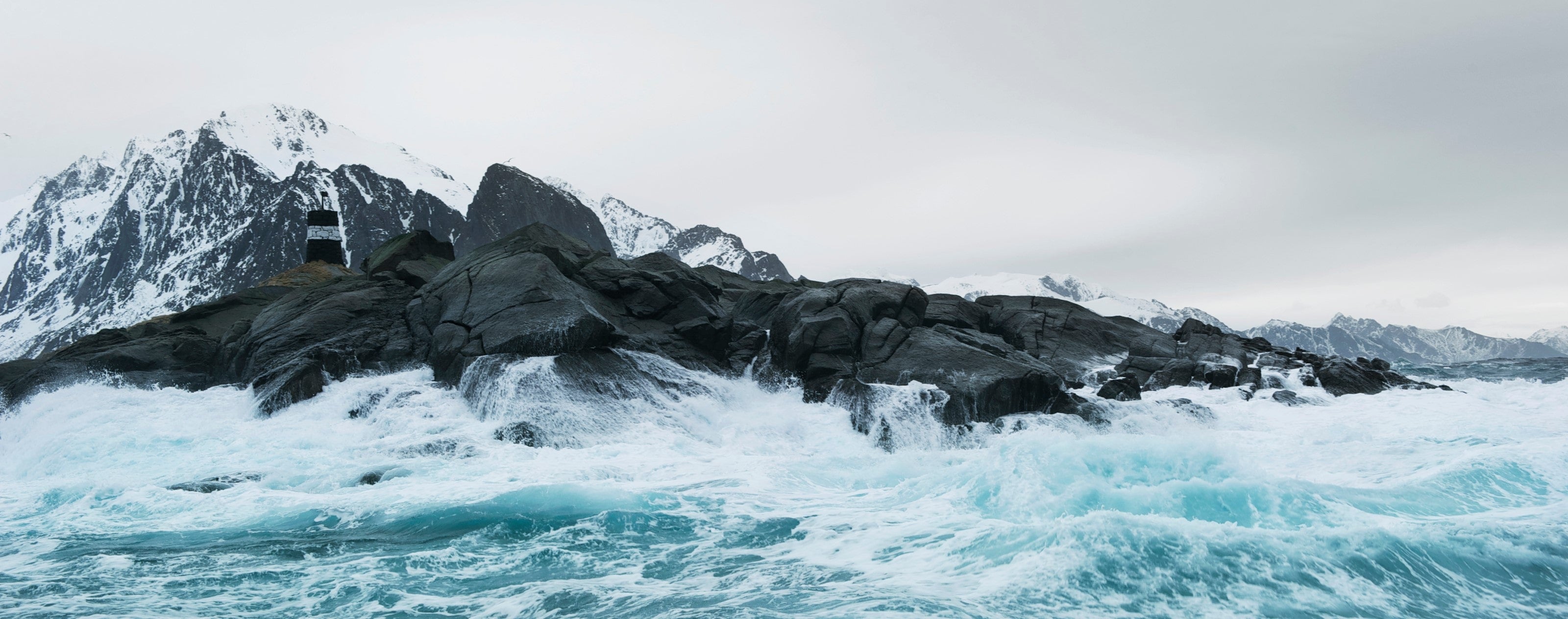 A rugged coastal landscape with powerful turquoise waves crashing against dark, jagged rocks. In the background, snow-covered mountains rise under an overcast sky. A small black-and-white navigation marker stands on the rocky terrain, contrasting with the wild natural surroundings. The scene captures the raw beauty and harsh conditions of an Arctic or Nordic coastline.
