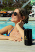 Woman relaxing by a pool in the sun, wearing sunglasses and a blue swimsuit, smiling with a black Ballstad-branded container placed in front of her on the poolside deck.