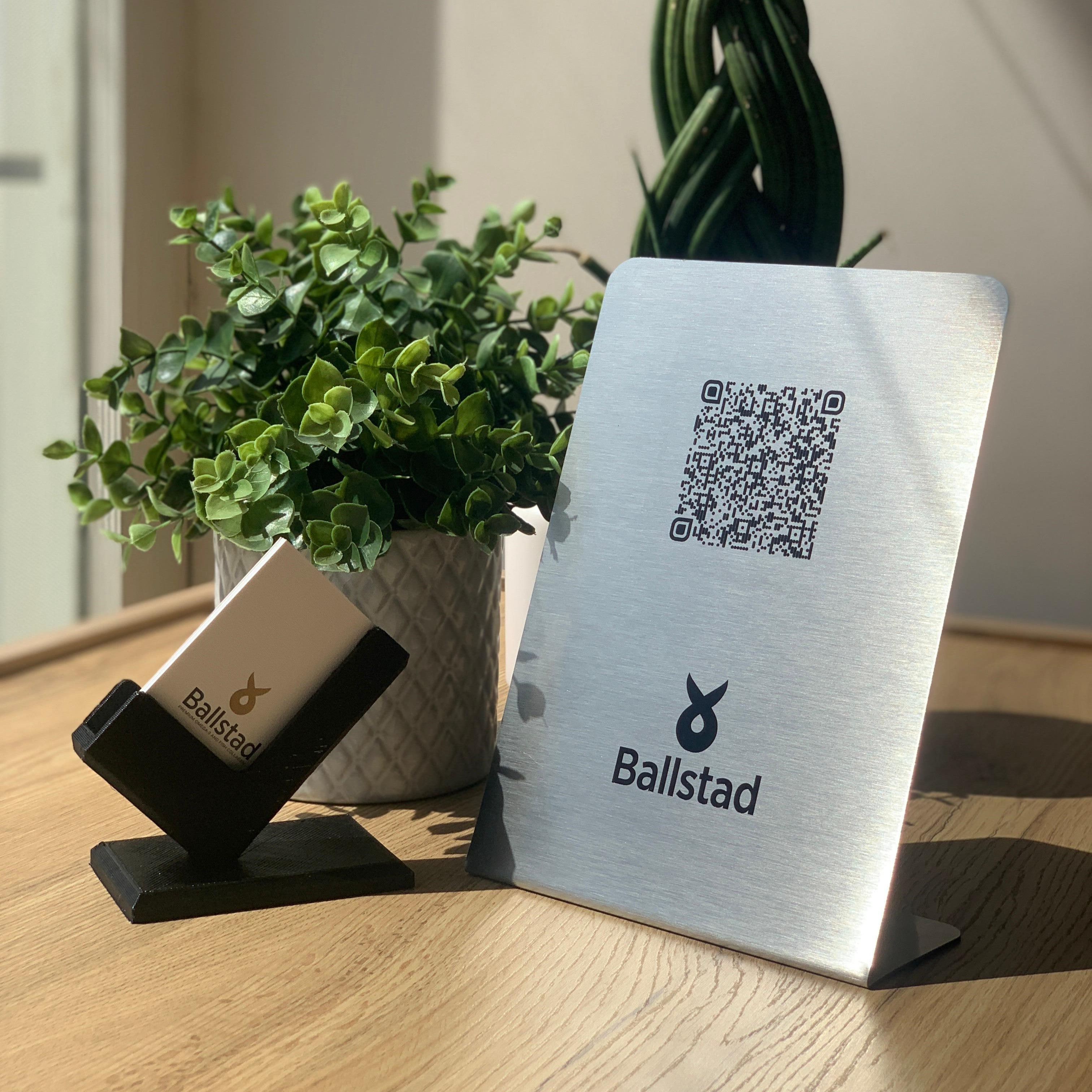 A sleek, brushed metal Ballstad QR code stand placed on a wooden surface, illuminated by natural sunlight. A small Ballstad-branded cardholder with a business card is positioned beside it, with a green potted plant in the background.