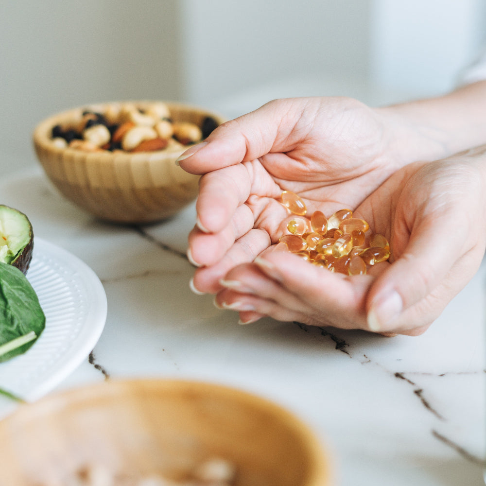 Close-up of a person's hands holding yellow omega-3 softgel capsules, with bowls of nuts, avocado, and leafy greens on a marble countertop in the background, symbolizing nutritional supplements and a healthy diet.