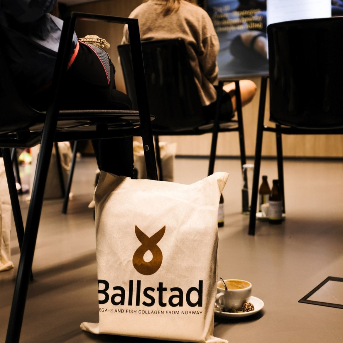 A Ballstad-branded tote bag made of natural fabric sits on the floor in a training or seminar setting. The bag features the Ballstad logo and the text 'Omega-3 and Fish Collagen from Norway.' In the background, people are seated on black chairs, and a cup of coffee with a small treat rests on a saucer nearby.