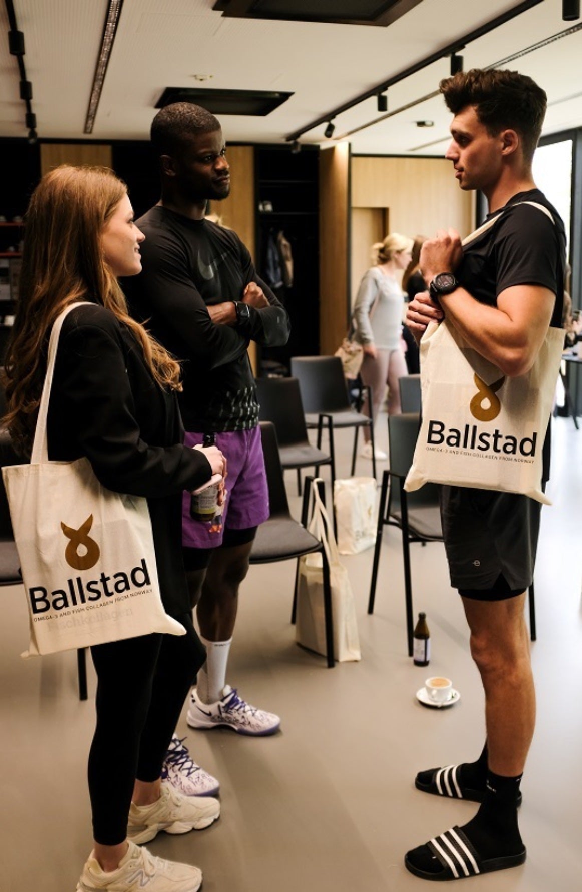 Three people chatting indoors, all holding or wearing Ballstad-branded tote bags. They appear to be in a casual setting, possibly at a wellness or fitness event, with chairs, drinks, and other Ballstad products in the background.