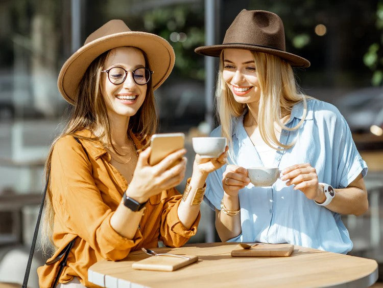Two stylish young women wearing hats and enjoying coffee at an outdoor café, smiling and looking at something on a smartphone together.
