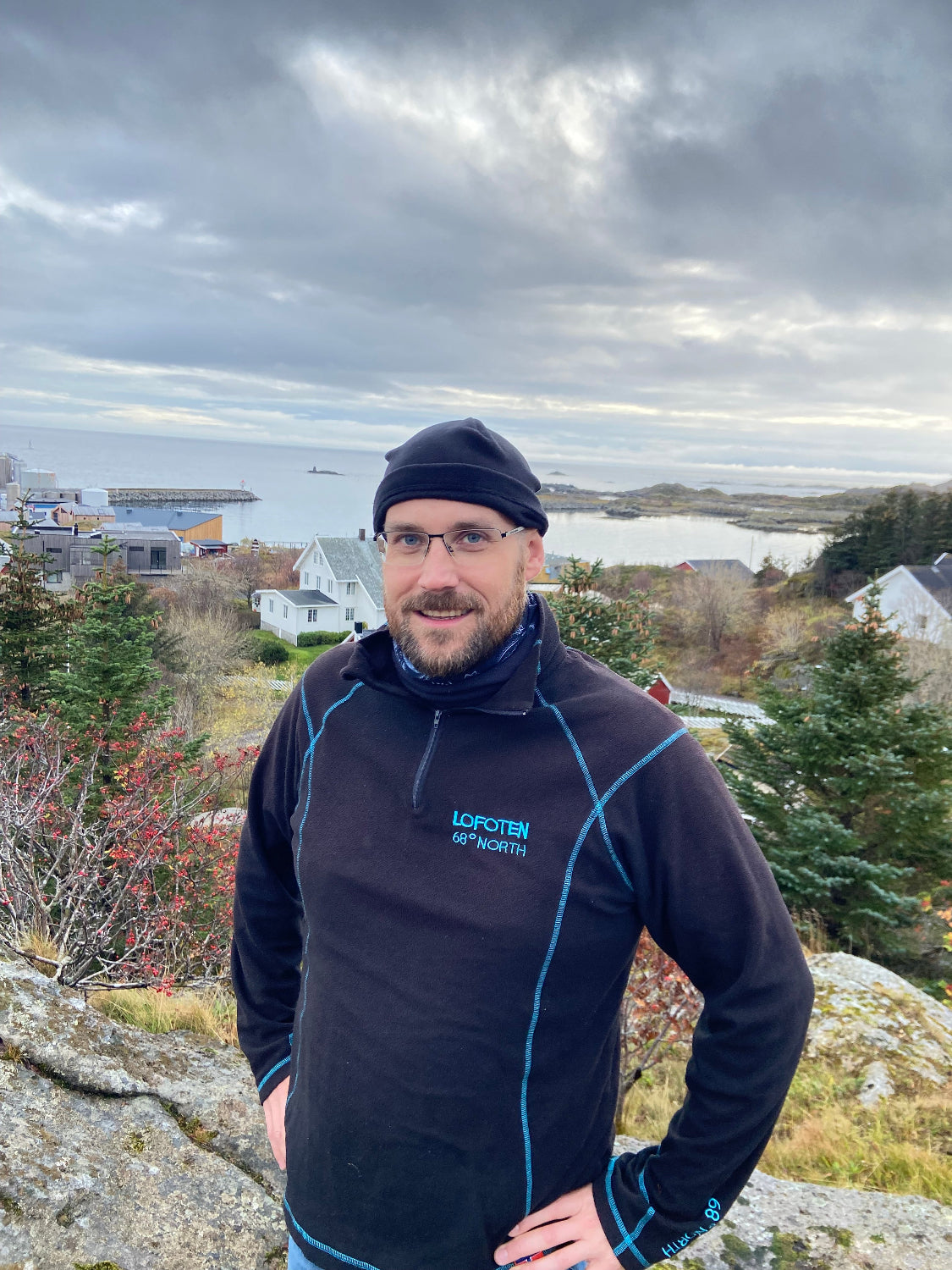 En mann med briller og svart lue står utendørs i Lofoten med utsikt mot havet og fjellene i bakgrunnen. Han har på seg en svart genser med blå detaljer og teksten "LOFOTEN 68° NORTH".