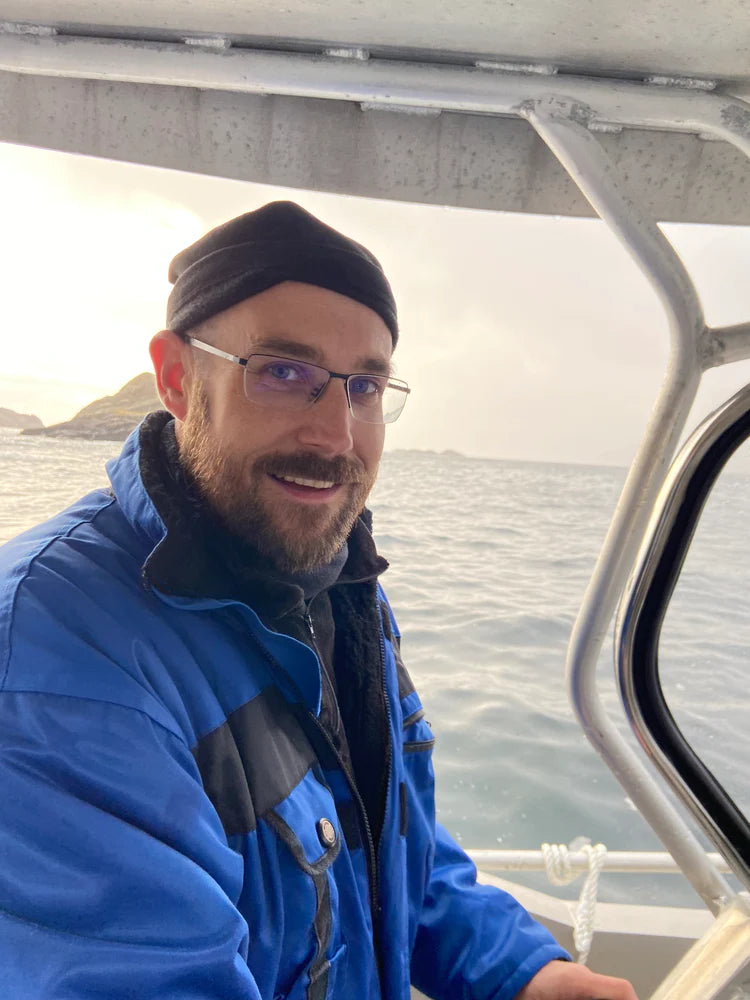 A man with a beard, glasses, and a black beanie is smiling while steering a boat. He is wearing a blue and black waterproof jacket with a black fleece underneath. The background features an open sea with small islands visible in the distance under an overcast sky. The boat has a metal frame and a covered roof.
