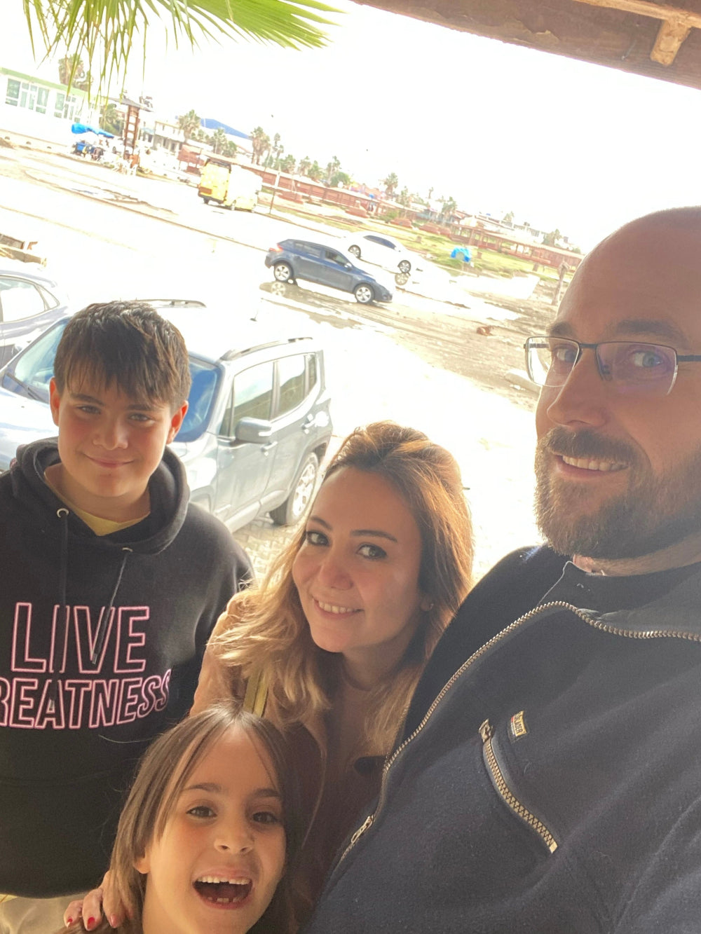 Happy family of four smiling outdoors near the coast — father in glasses, mother with blonde wavy hair, teenage boy in 'Live Greatness' hoodie, and cheerful young girl in front. Casual family photo with parked cars, palm trees, and seaside setting in the background.
