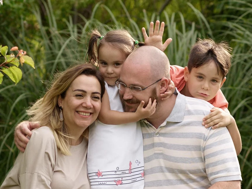 Family portrait of a joyful family of four, with two parents and two children embracing outdoors in a lush, green setting. The mother smiles warmly on the left, the father is in the center with one child hugging him from behind, and the second child stands close by, all sharing a moment of togetherness and affection.