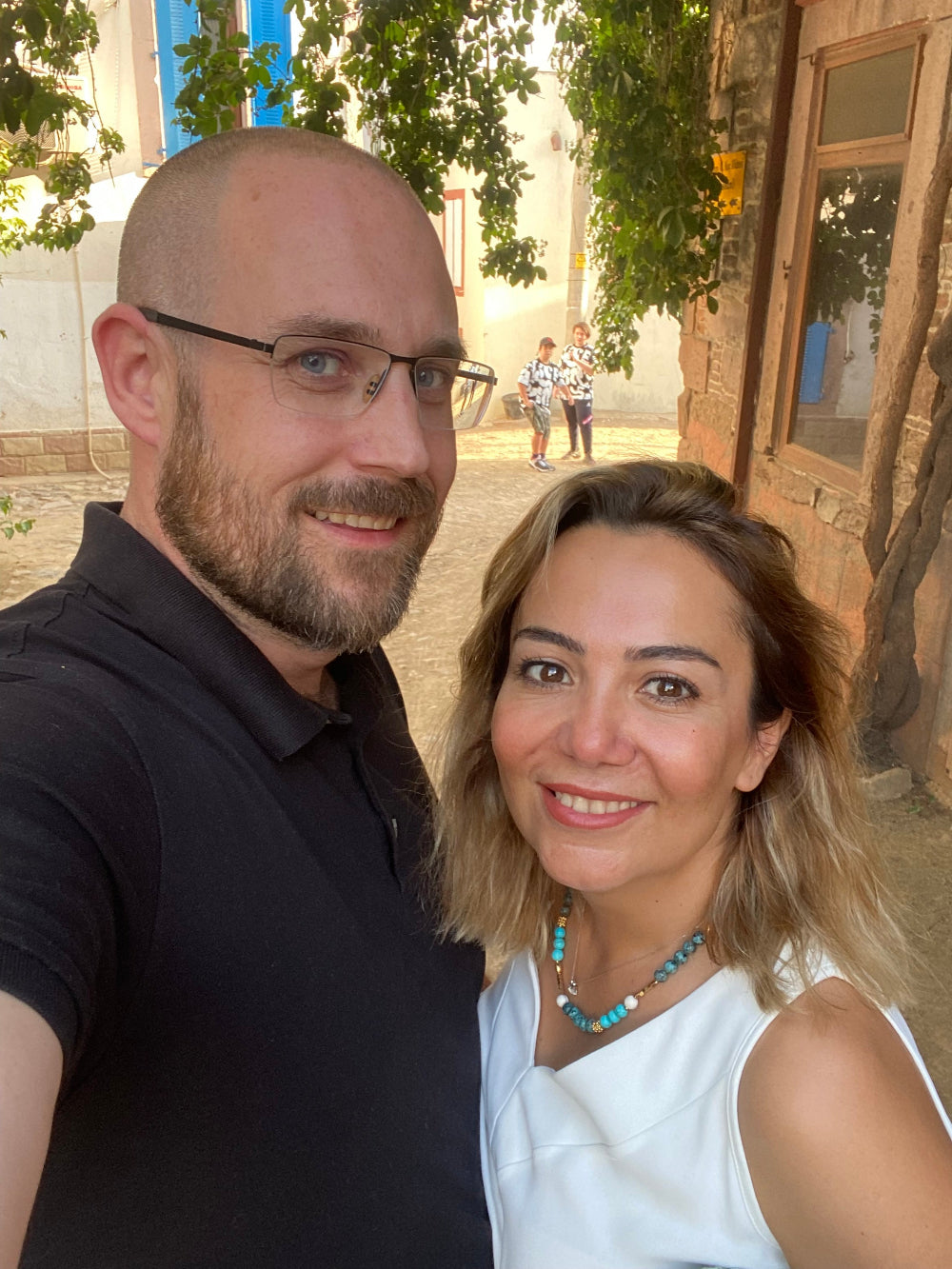 A smiling couple takes a selfie outdoors in a charming, sunlit alley with stone walls and green foliage overhead. The man has a beard, glasses, and wears a black polo shirt, while the woman has shoulder-length blonde hair, wears a white sleeveless top, and a turquoise necklace. In the background, two children in matching outfits are walking.