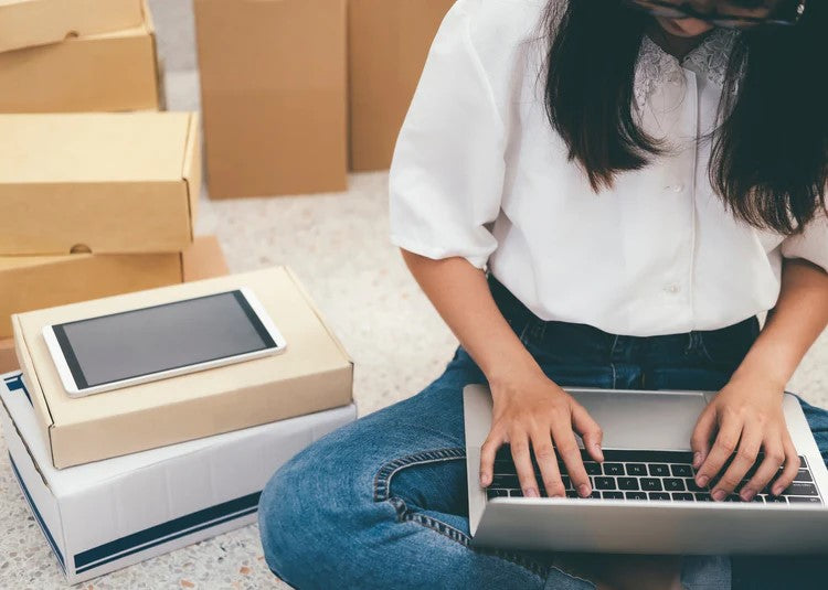 A woman sitting on the floor working on a laptop, surrounded by cardboard boxes, with a tablet placed on top of one of the boxes—suggesting she is managing an online business or handling e-commerce orders.