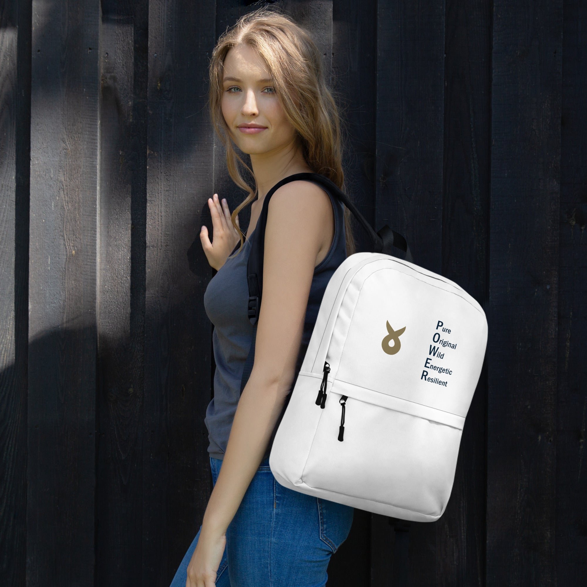 Young woman with long blonde hair, wearing a dark tank top and jeans, stands against a dark wooden wall, carrying a white backpack featuring the Ballstad logo and the acronym "POWER" vertically spelled as Pure, Original, Wild, Energetic, Resilient.
