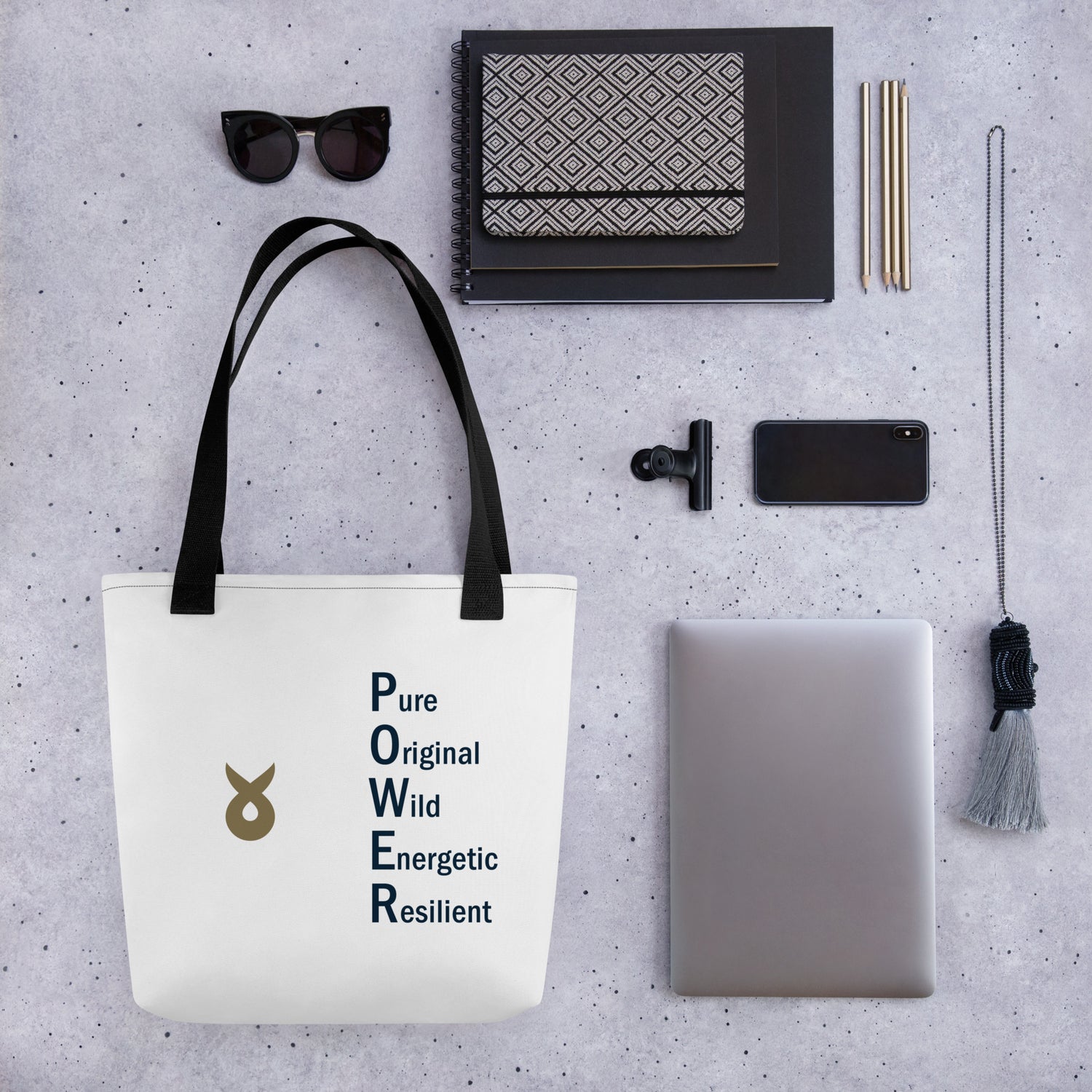 A white tote bag with black handles lies on a speckled grey surface, featuring the Ballstad logo and the acronym “POWER” spelled vertically with words Pure, Original, Wild, Energetic, Resilient. Surrounding the bag are neatly arranged accessories including sunglasses, notebooks, pencils, a phone, a laptop, and a decorative tassel.