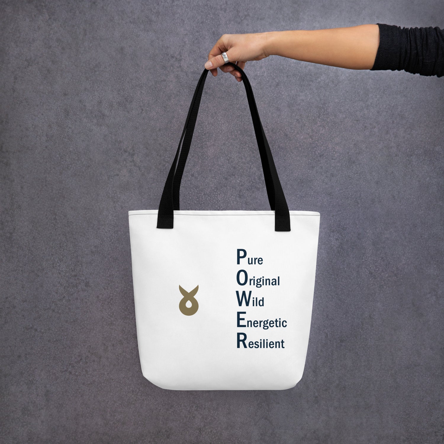 A person holding a white tote bag by its black straps against a grey textured background. The bag features the Ballstad logo and the word "POWER" vertically spelled out with accompanying words: Pure, Original, Wild, Energetic, Resilient.