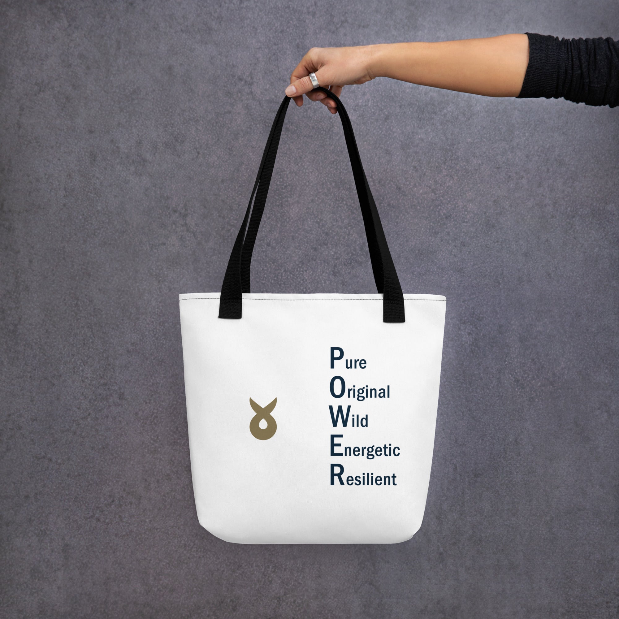 A person holding a white tote bag by its black straps against a grey textured background. The bag features the Ballstad logo and the word "POWER" vertically spelled out with accompanying words: Pure, Original, Wild, Energetic, Resilient.