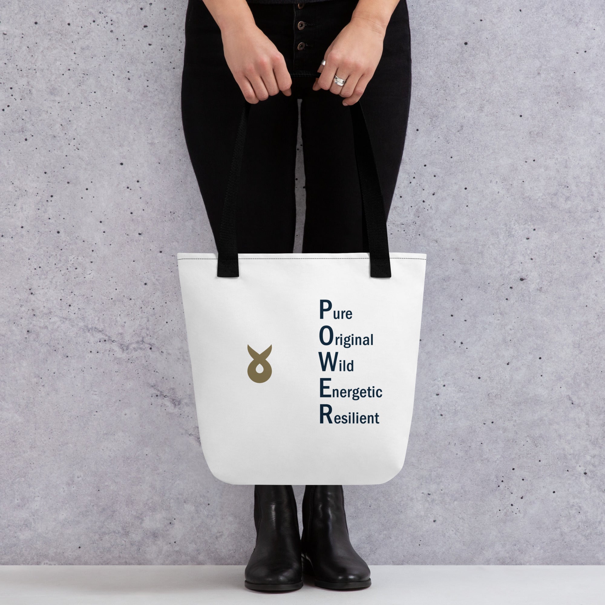 A person dressed in black pants and black boots is holding a white tote bag with black straps. The bag features the Ballstad logo and the acronym “POWER” spelled vertically with the words “Pure, Original, Wild, Energetic, Resilient” printed beside each corresponding letter. The background is a light gray concrete wall.