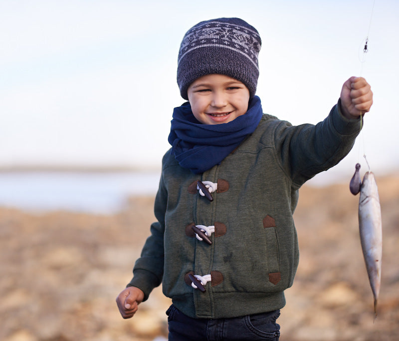 Smiling young boy wearing a winter hat and scarf, proudly holding a freshly caught fish by the shoreline.