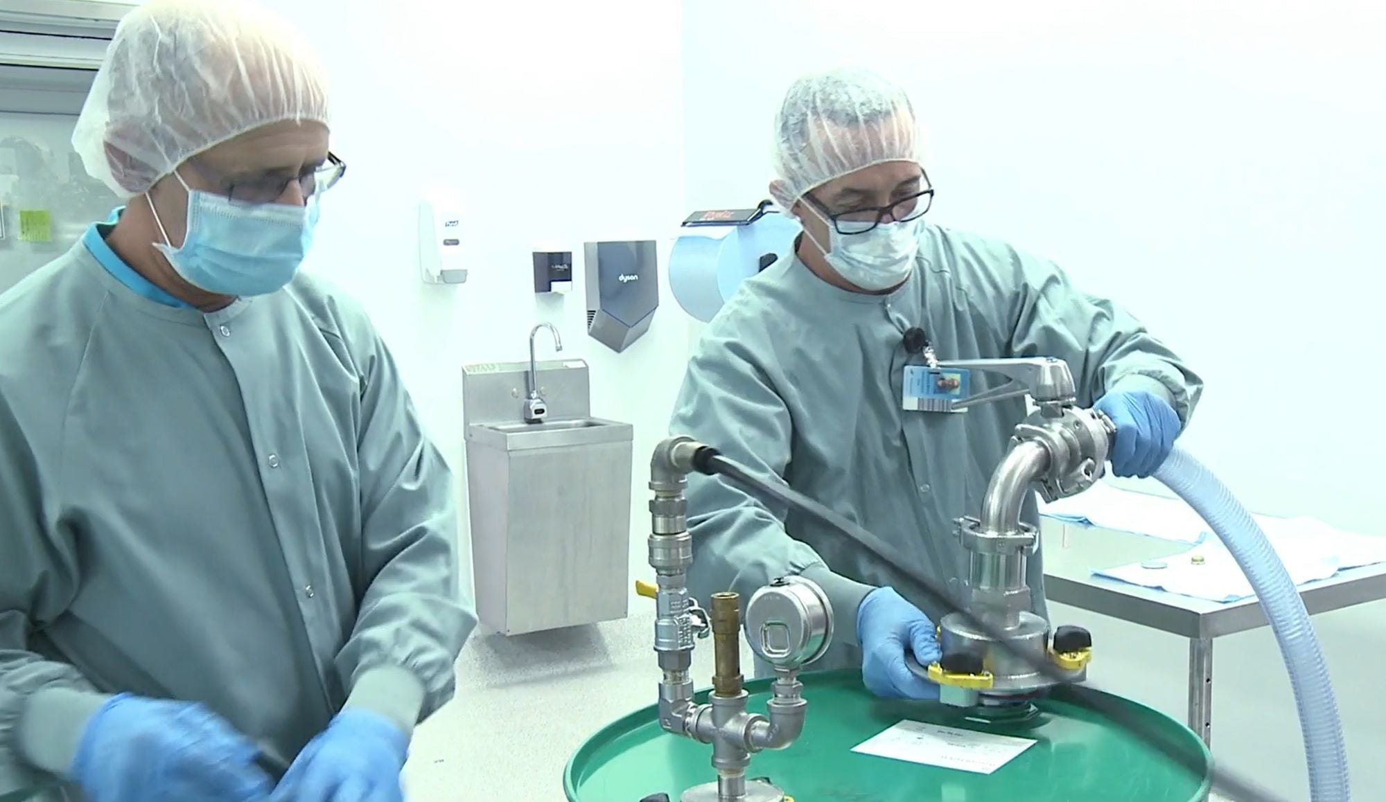 Two workers in a cleanroom environment are handling industrial equipment. They are wearing protective clothing, including gray lab coats, hairnets, face masks, safety glasses, and blue gloves. One worker is operating a metal hose connected to a green drum, while the other is assisting. The background features a stainless steel sink, wall-mounted dispensers, and a clean workspace, suggesting a sterile or pharmaceutical manufacturing setting.