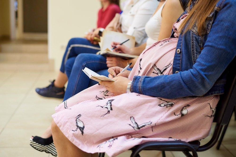 Pregnant woman in a pink dress with bird print taking notes while seated in a waiting room with other women.