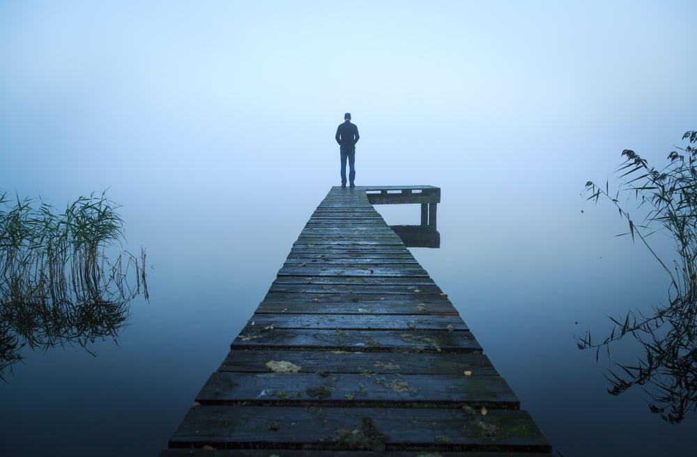 Silhouetted person standing alone at the end of a wooden dock surrounded by calm water and dense fog, evoking solitude, reflection, or emotional isolation.