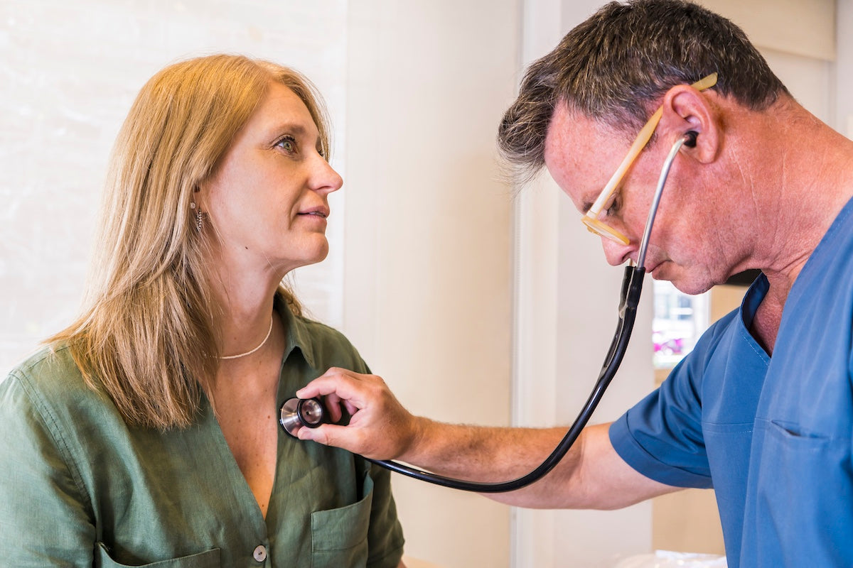 A mid-adult doctor wearing scrub clothes checking her patient's heart beat with stethoscope.