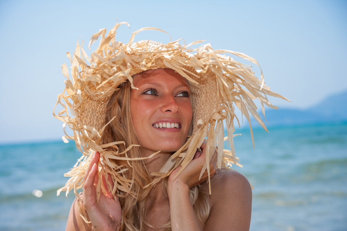 Smiling woman wearing a straw hat enjoying a sunny day at the beach with the sea in the background.