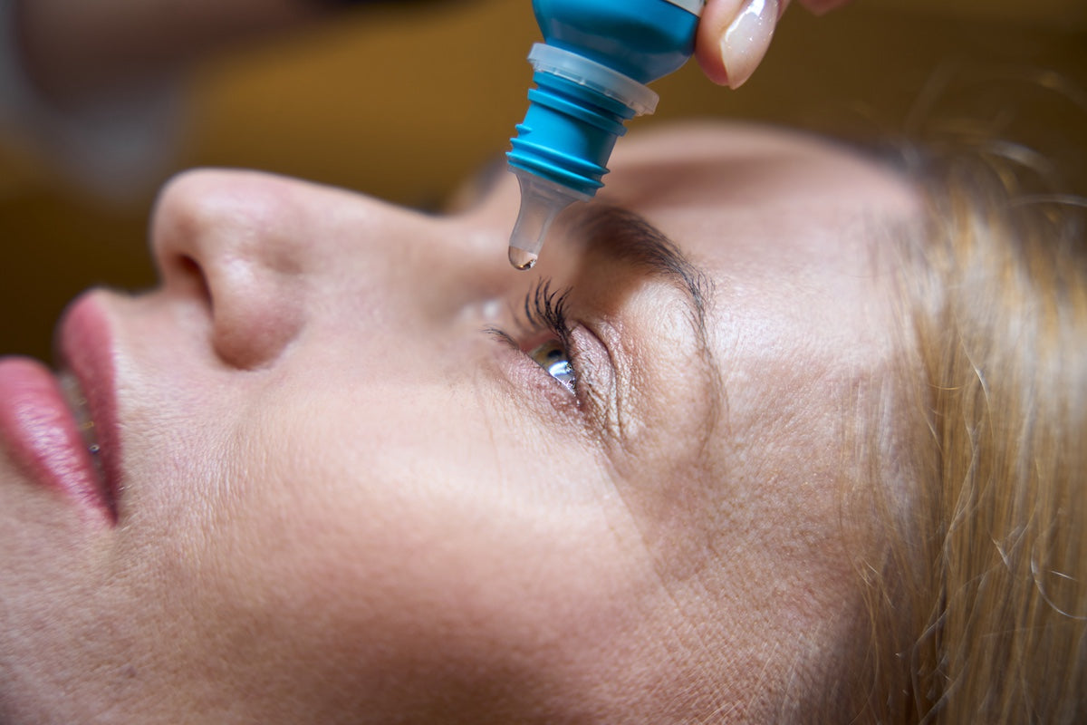 A blonde woman lying down receives an eye drop from a blue-capped bottle into her open eye.