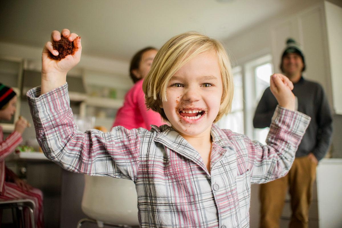 Smiling young child with blonde hair and chocolate on their face holds up a piece of cake in a kitchen, celebrating joyfully while family members look on in the background.