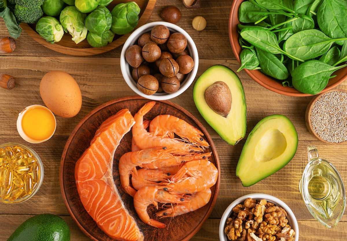 An assortment of omega-3-rich foods arranged on a wooden table, including salmon, shrimp, avocado, walnuts, macadamia nuts, spinach, Brussels sprouts, broccoli, eggs, fish oil capsules, and a glass container of oil.