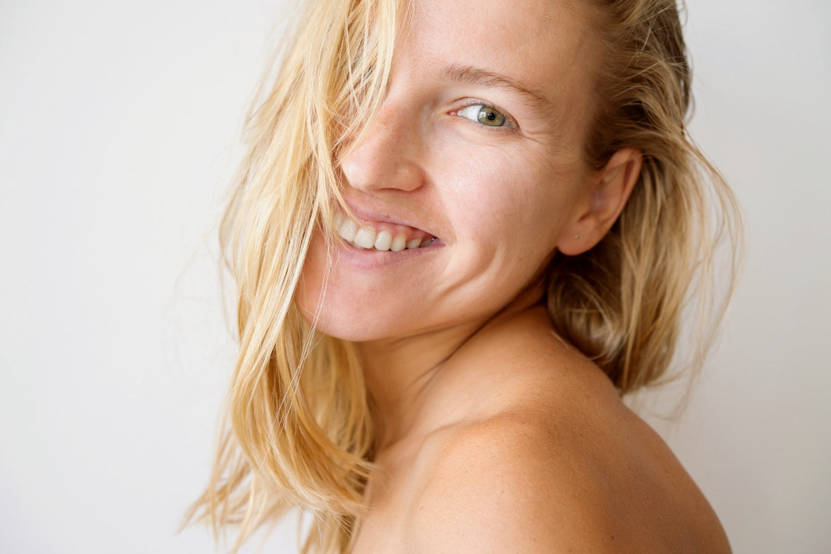 A young woman with blonde hair, healthy skin, and a happy expression is smiling against a white background; reflecting the theme of skin health and natural beauty.